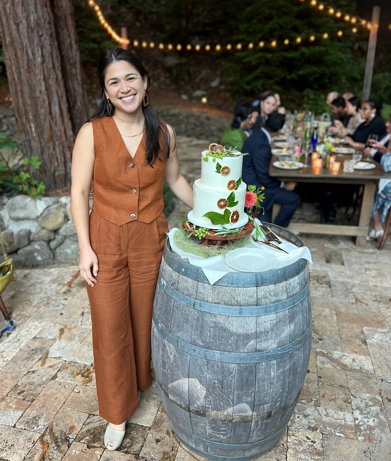 Baker standing next to a two-tier citrus-decorated wedding cake at an outdoor celebration